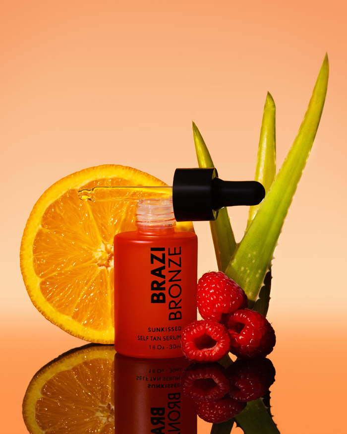 A bottle of Brazil Bronze Sunkissed Self Tan Serum sits among an orange slice, raspberries, and aloe vera on a reflective orange background.