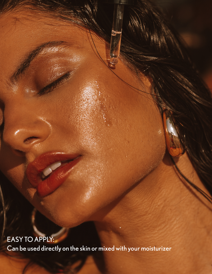 A woman with glowing, sun-kissed skin applies a drop of hydrating facial serum directly to her cheek from a glass dropper.