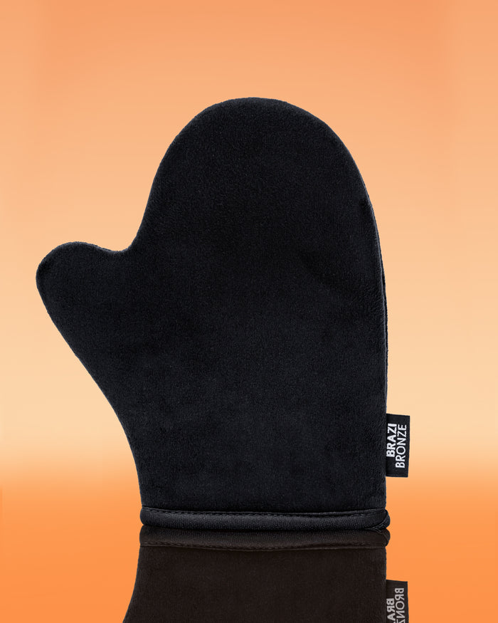 A plush black velvet Brazi Bronze tanning mitt stands upright on a reflective surface against a warm orange background for smooth sunless tan application.